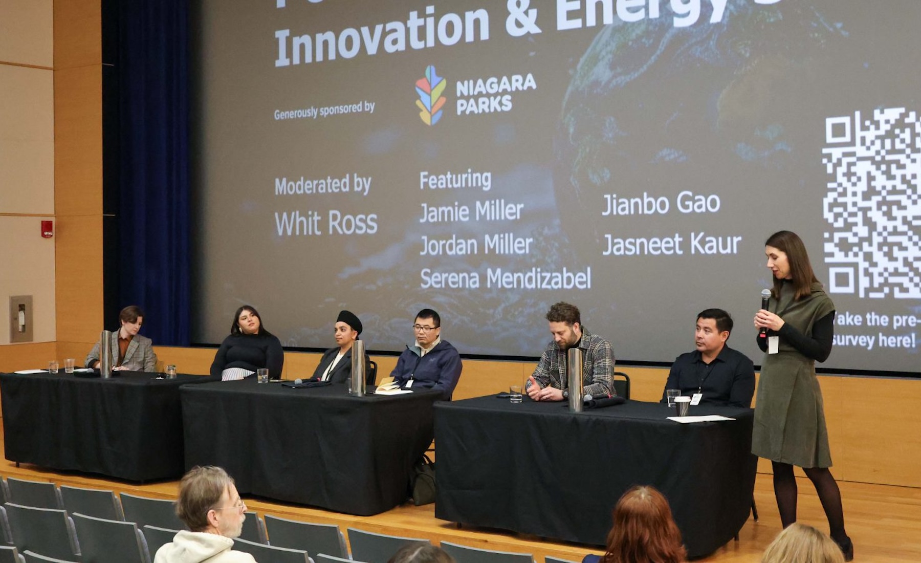 A group of people sit at a table on stage during a panel while a woman holding a microphone stands nearby. 