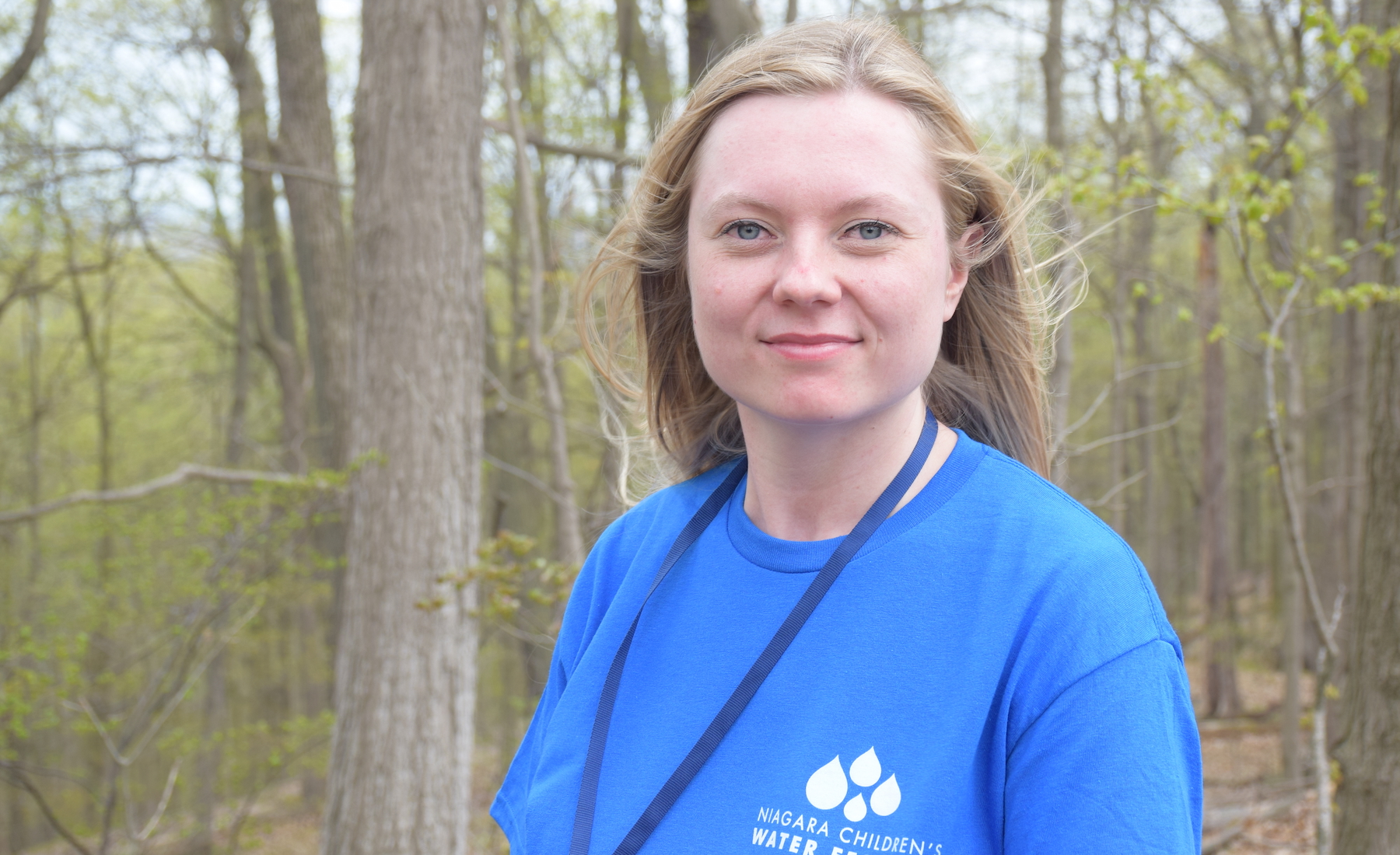 Brock grad Kassie Burns (BSc ’22, MS ’24) stands in front of a group of trees wearing a Niagara Children's Water Festival shirt.