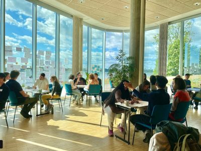 A group of people sitting at desks in a brightly lit room with floor to ceiling windows.