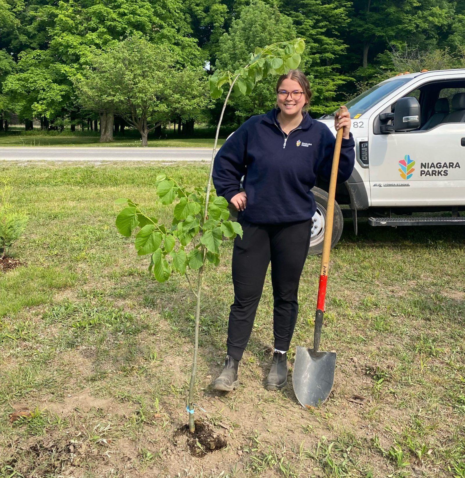 Student standing by a newly planted tree holding a shovel with a Niagara Parks truck in the background.