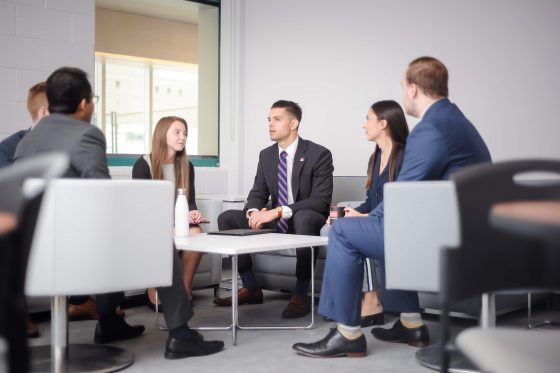 "A group of six professionally dressed individuals sit in a casual meeting area, engaged in conversation around a low white table in a modern office setting."