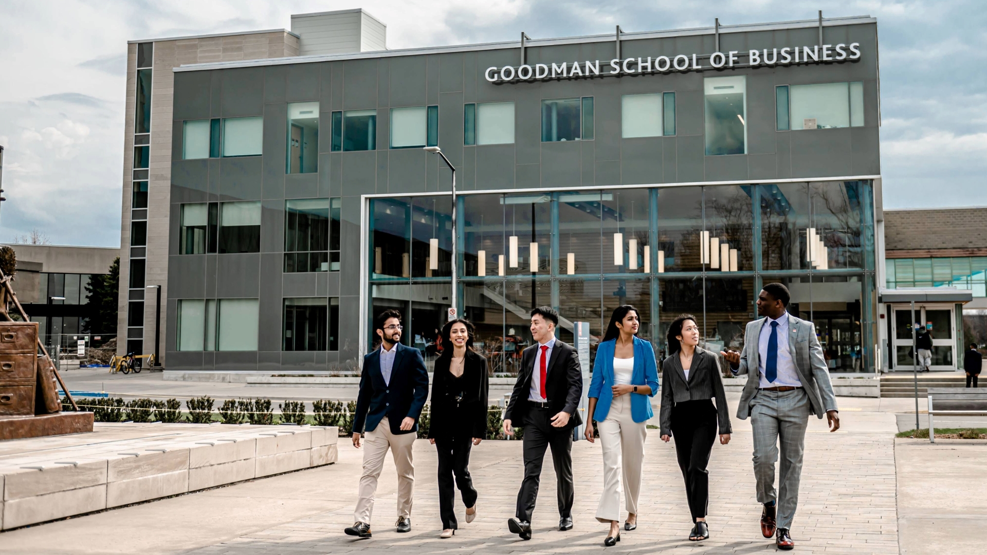 Six business students walk in front of the Goodman School of Business building. 