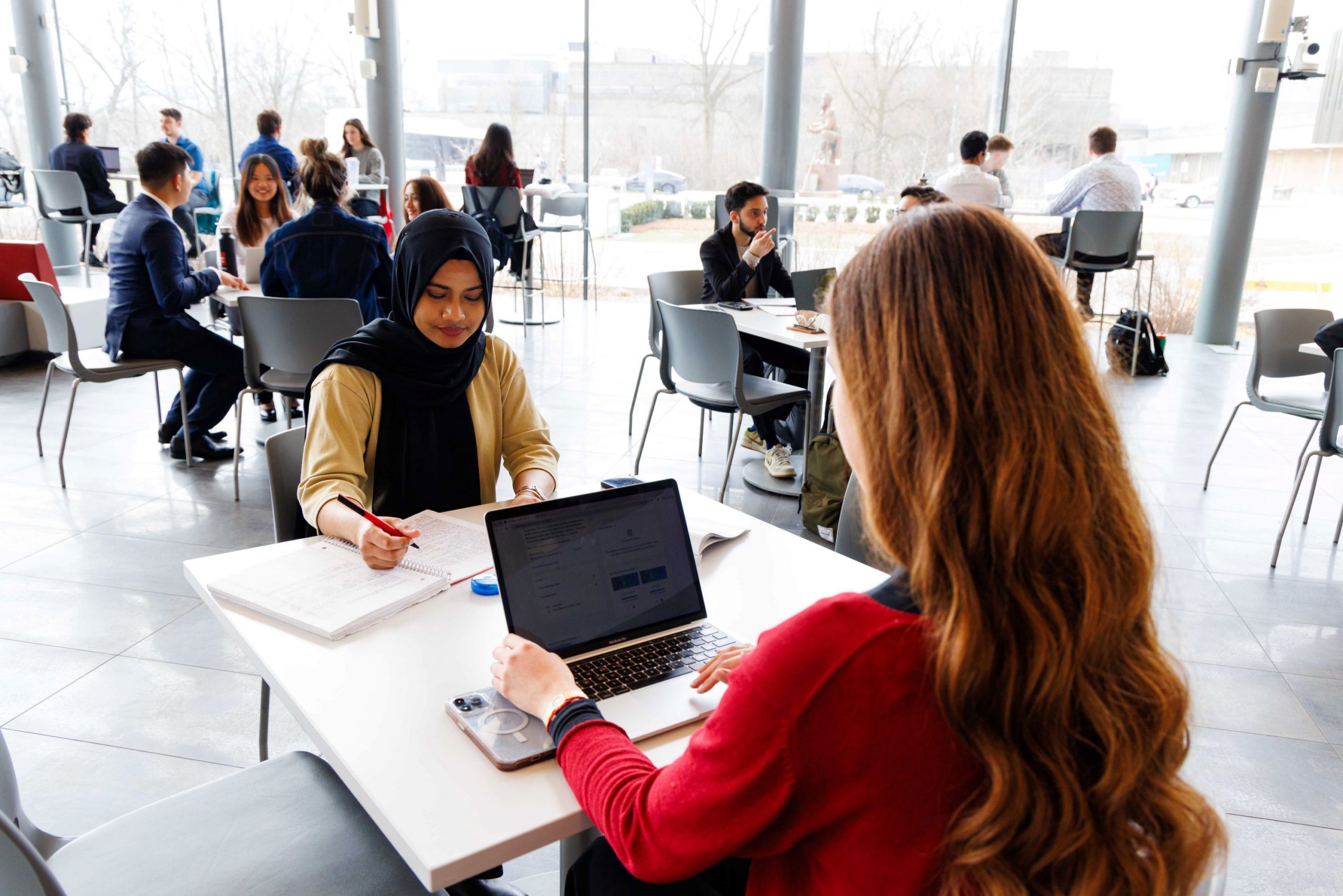 Two students sit across from each other at a table, one writing in a notebook while the other types on her laptop, surrounded by other students engaging in conversation in the background