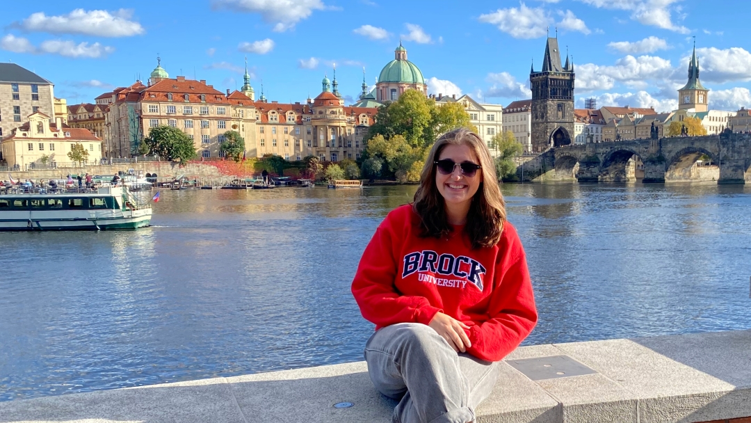 Student sits in front of a canal in Europe with a city landscape in the background. 