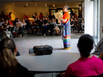A women sings and plays a hand drum in the centre of a room full of onlookers.