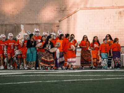 A group of young women sing and play hand drums while leading male lacrosse players onto the field.