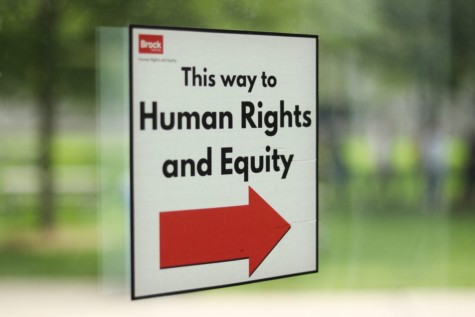 A printed sign taped to a glass window or door. The sign has a white background and says 'This way to Human Rights and Equity' in bold black text. Below the text is a large red arrow pointing to the right. The Brock University logo is displayed in the top left corner of the sign.