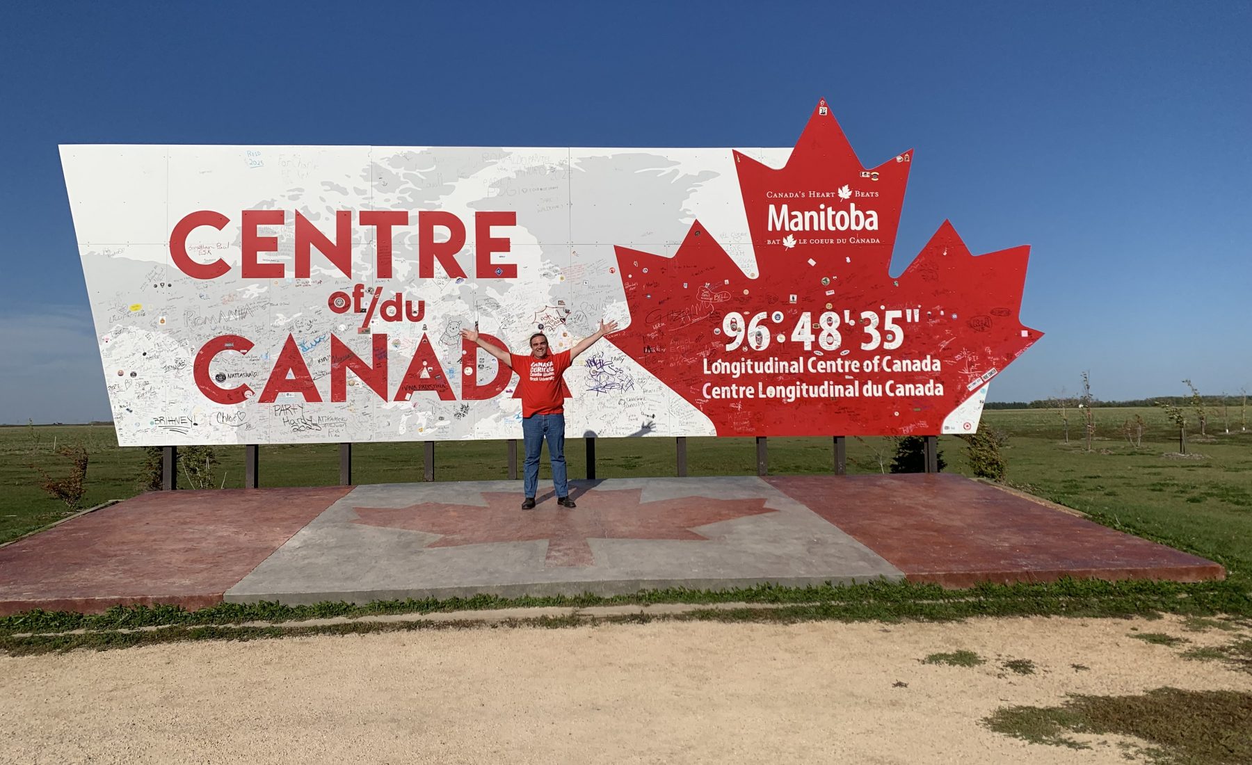A man stands with arms outstretched in front of a large sign marking the Longitudinal Centre of Canada in Manitoba, located at 96° 48' 35