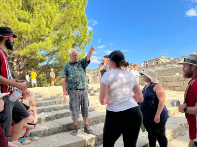 6 students look at professor pointing sitting on the steps of the Theatral Area at Knossos