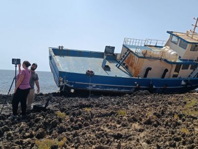 3 students at a camera tripod next to a large blue boat on shore