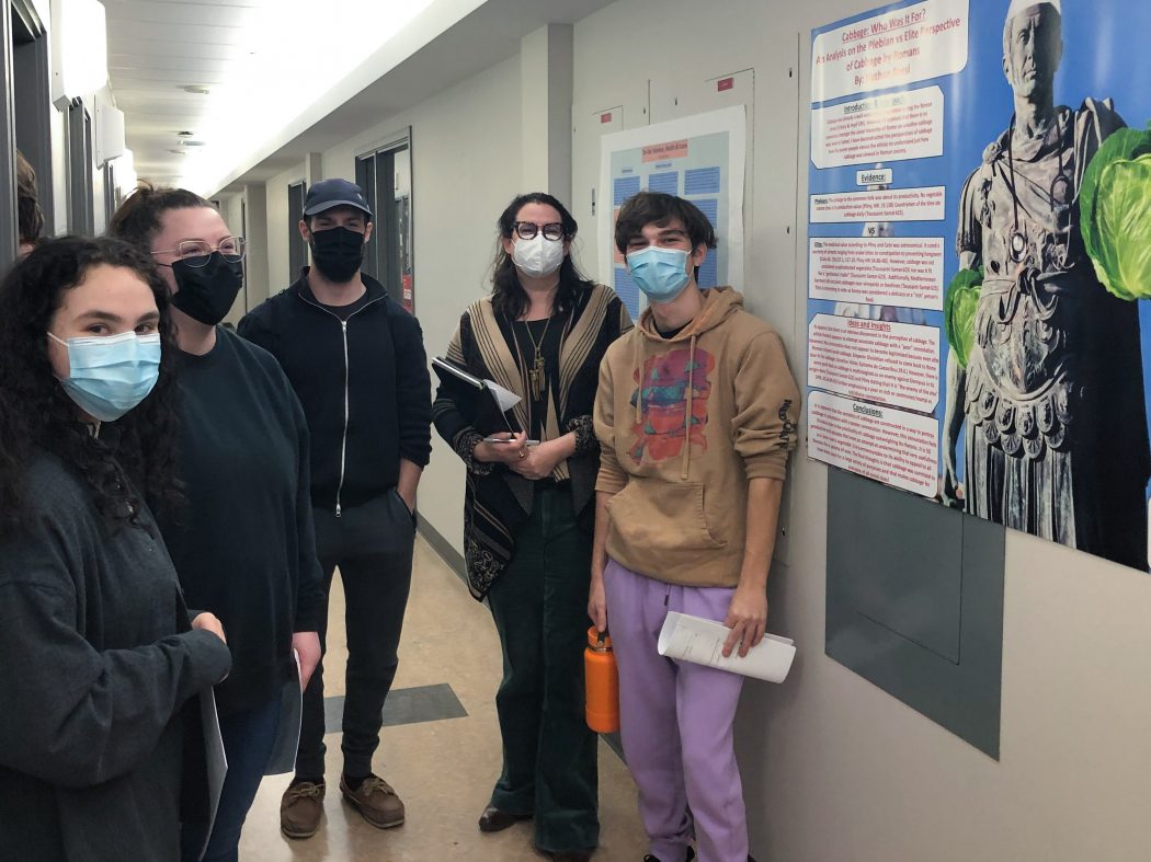 5 students in covid masks in a hallway with a poster of Alexander the great