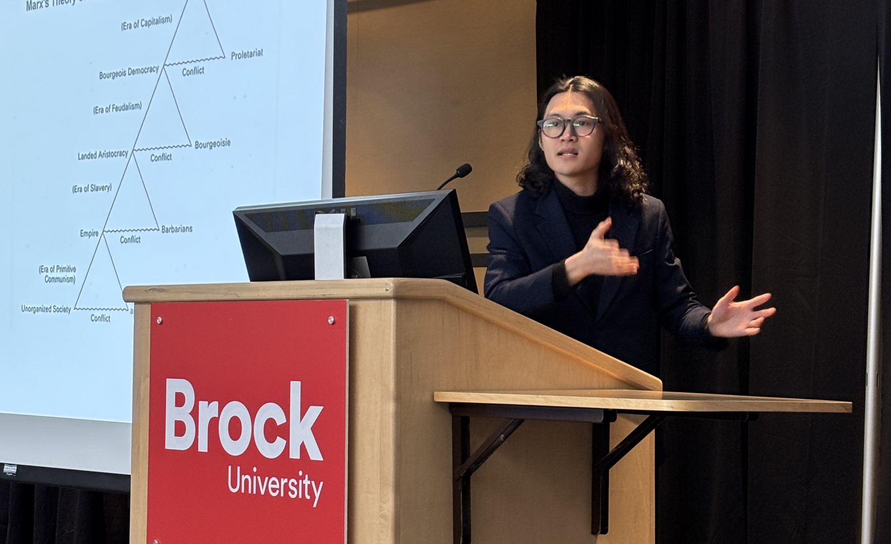 A speaker gestures during a presentation at a Brock University lectern, with a diagram of Marx's Theory of History on the screen behind them. 