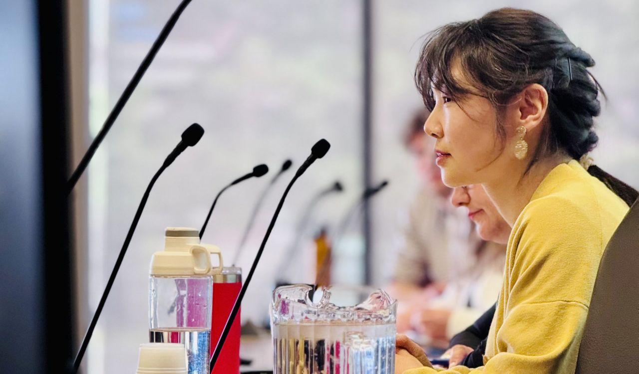 A woman in a yellow top seated at a conference table with microphones and water bottles, listening attentively during a meeting. 