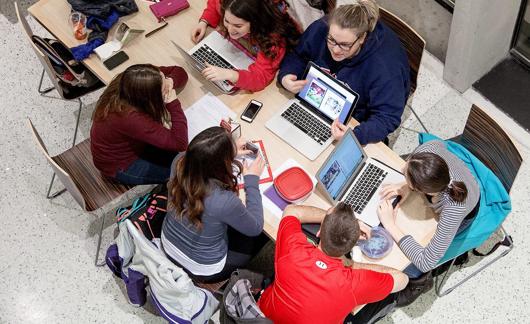 6 students from above sitting at table showing each other laptop screens