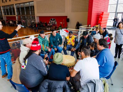 People sit in a circle preparing to hit a drum placed in the centre.