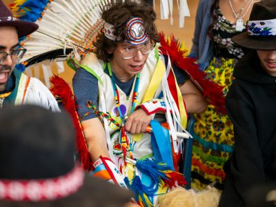 A group of men in traditional Indigenous regalia sitting in a gymnasium.