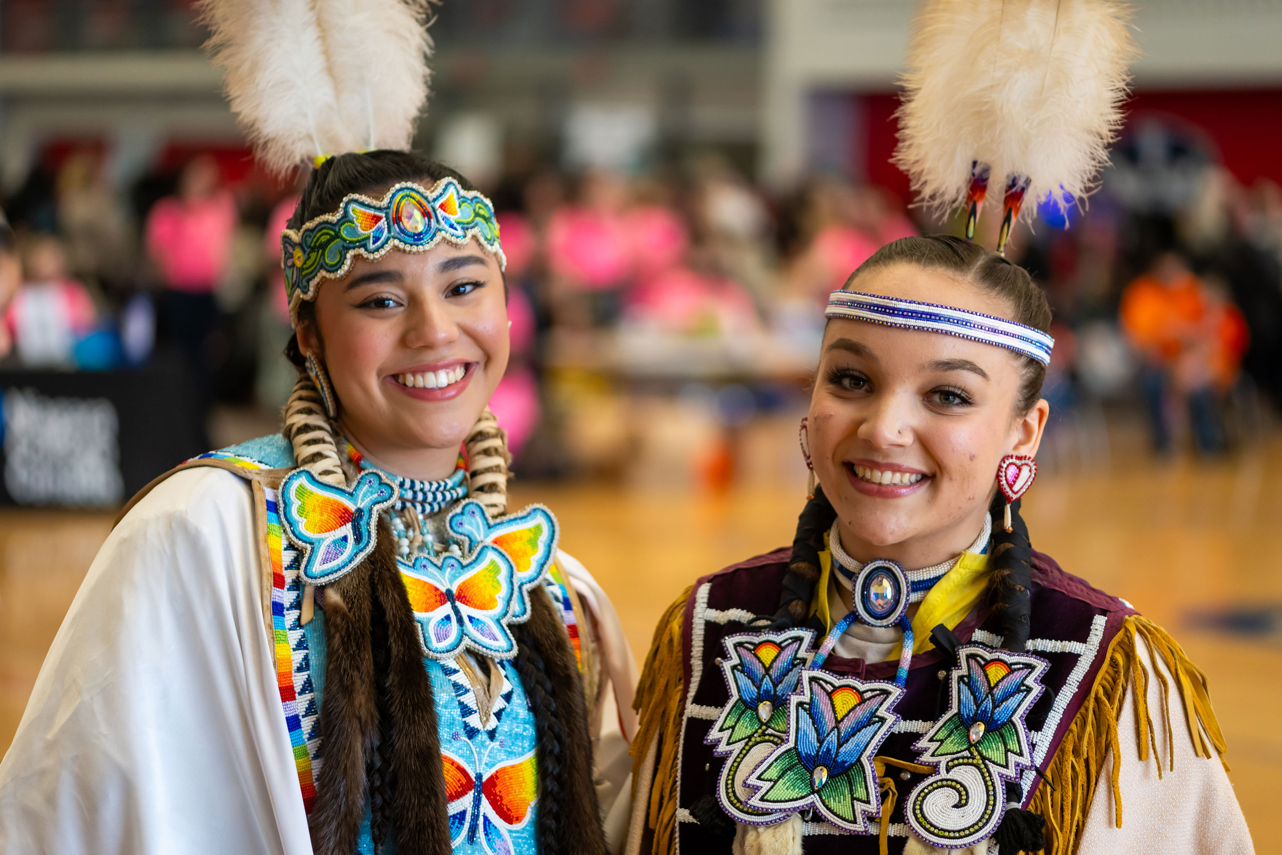 Two women standing together smiling and dressed in traditional Indigenous regalia.