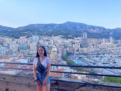 A woman smiles on a bridge overlooking buildings and mountains in Monaco.