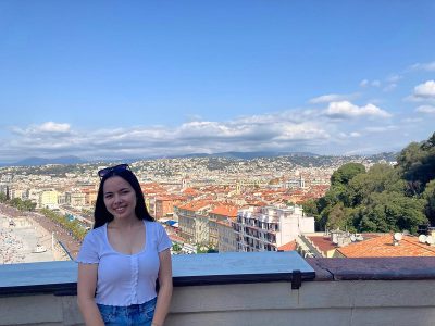 A woman smiles for a photo at a scenic lookout, looking down on buildings with orange rooftops.