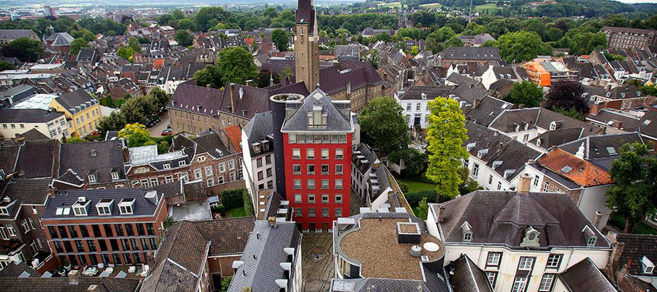 An aerial view of the European town of Maastricht with densely packed traditional buildings, featuring a prominent red building in the center and a tall clock tower in the background.