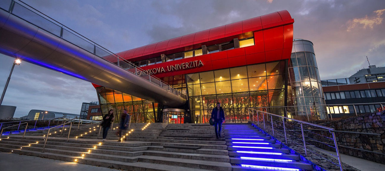 A university student walking up a blue LED-lit staircase towards a modern red and glass building at Masaryk University in the evening.