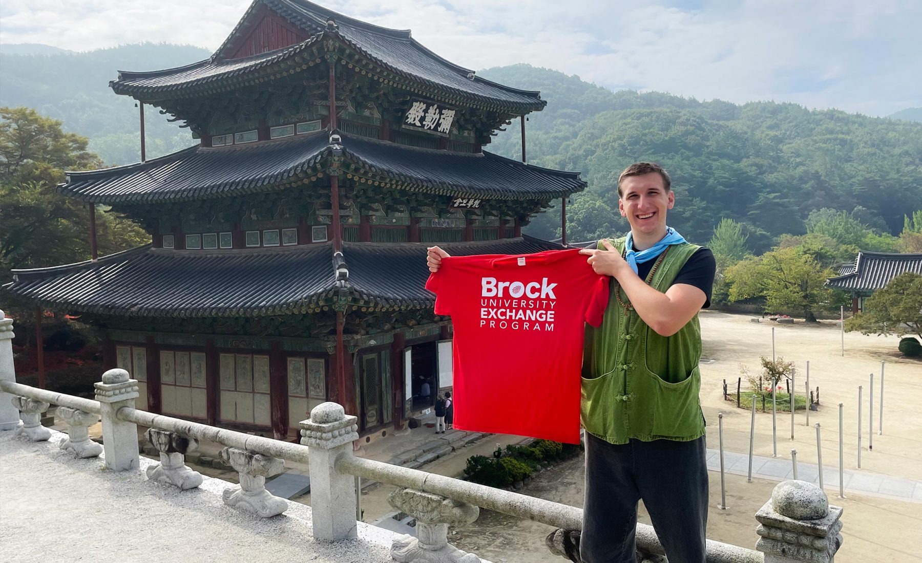 A university student on exchange holds a Brock University Exchange Program t-shirt while standing in front of a traditional Japanese building. 