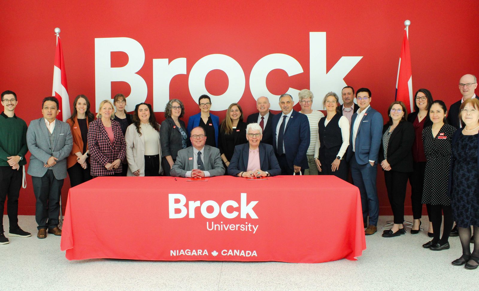 A large group of people stands behind two people signing a document at a long table with the flags of Canada and The Netherlands in the background. 