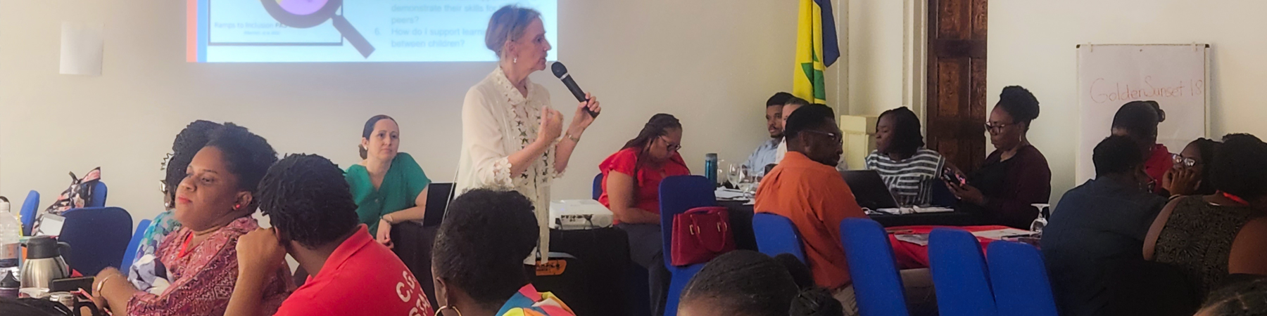 An instructor holds a microphone, speaking in the centre of a room with people working at tables around her.