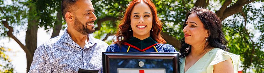 A Brock university graduate in a convocation robe holds a framed diploma as her parents stand on either side of her smiling at her.