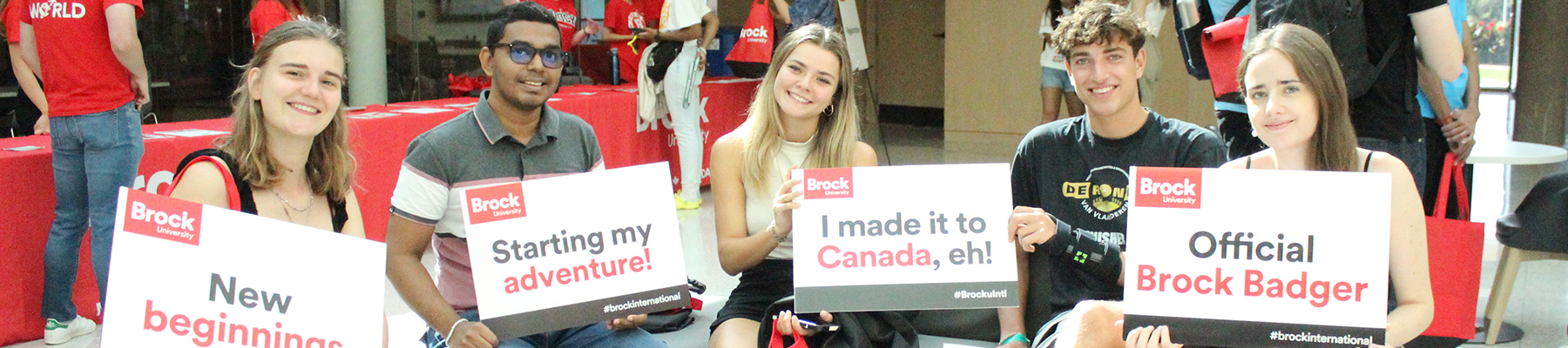 A group of university students hold signs about starting their adventure in Canada seated on a couch in a bright lobby area.