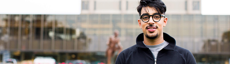 A man wearing glasses and a sweater smiles at the camera outdoors in front of a modern building on a cloudy day.