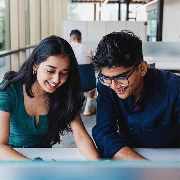 Two university students lean over a desk smiling at what they see in front of them.
