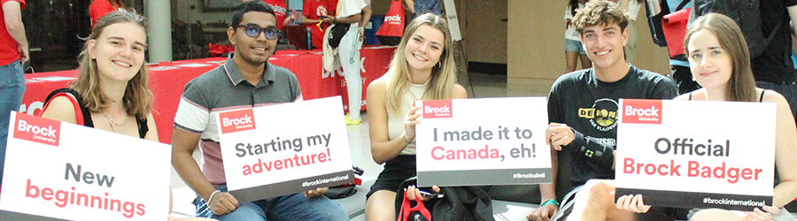 University students sit on a couch side-by-side holding signs referring to their first day in Canada and at university.