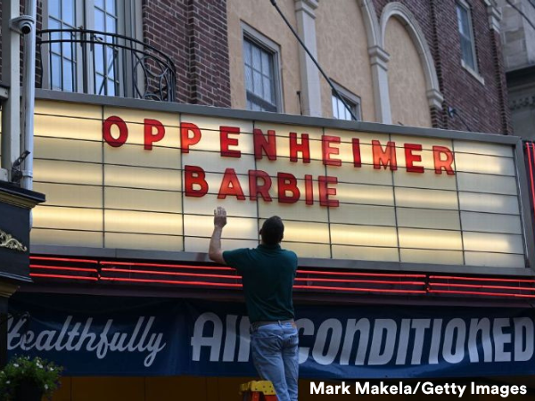 An illuminated movie theatre sign features the film names: Oppenheimer and Barbie. A sign installer atop a ladder works to install the letters.
