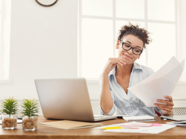 A woman works on a laptop and holds documents in her hand
