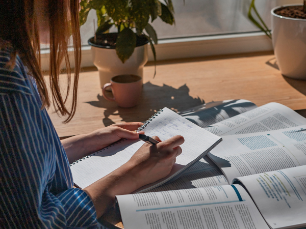 A woman works at a desk next to a window. Documents and highlighted books are spread out in front of her.