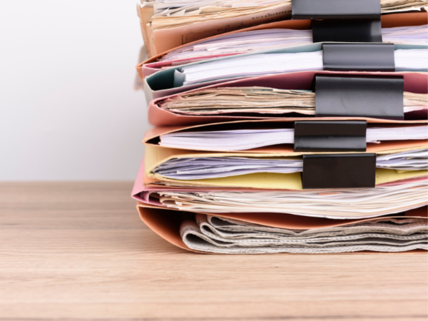A stack of manila file folders on a desk. Each folder is stuffed with documents.