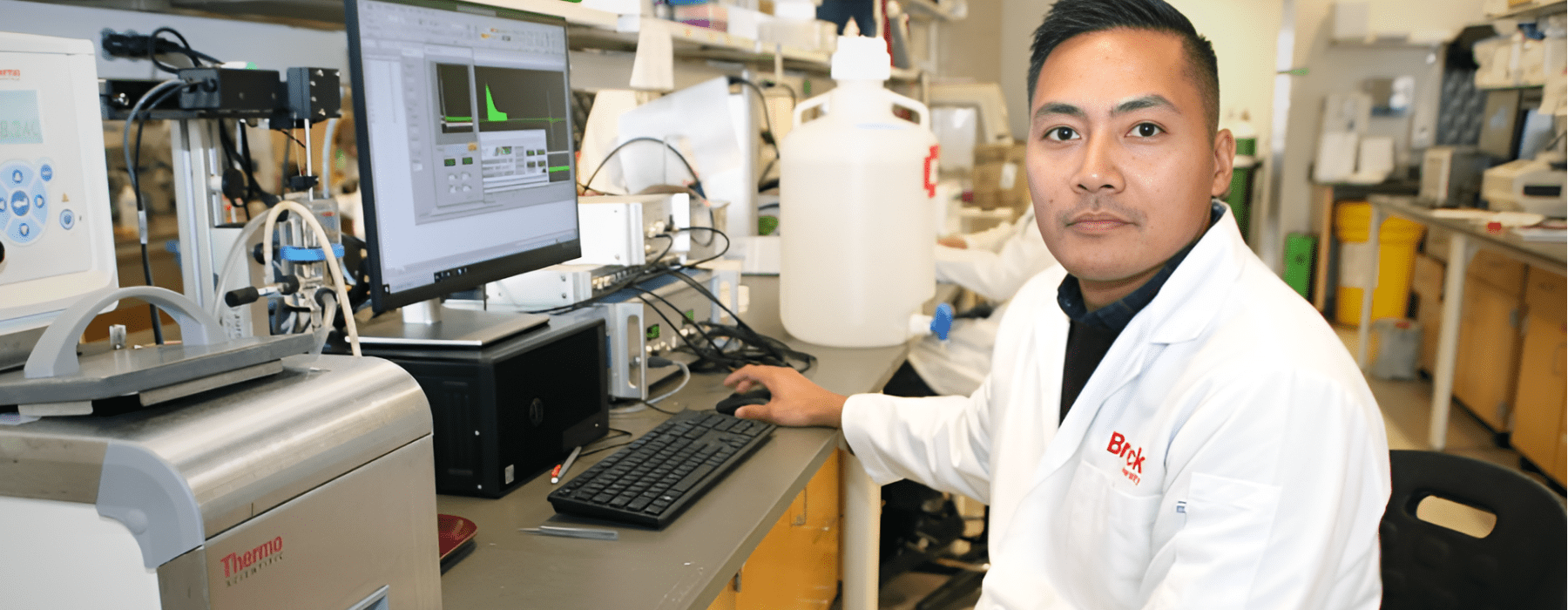 Student in a Brock University lab jacket in front of computer and research equipment 