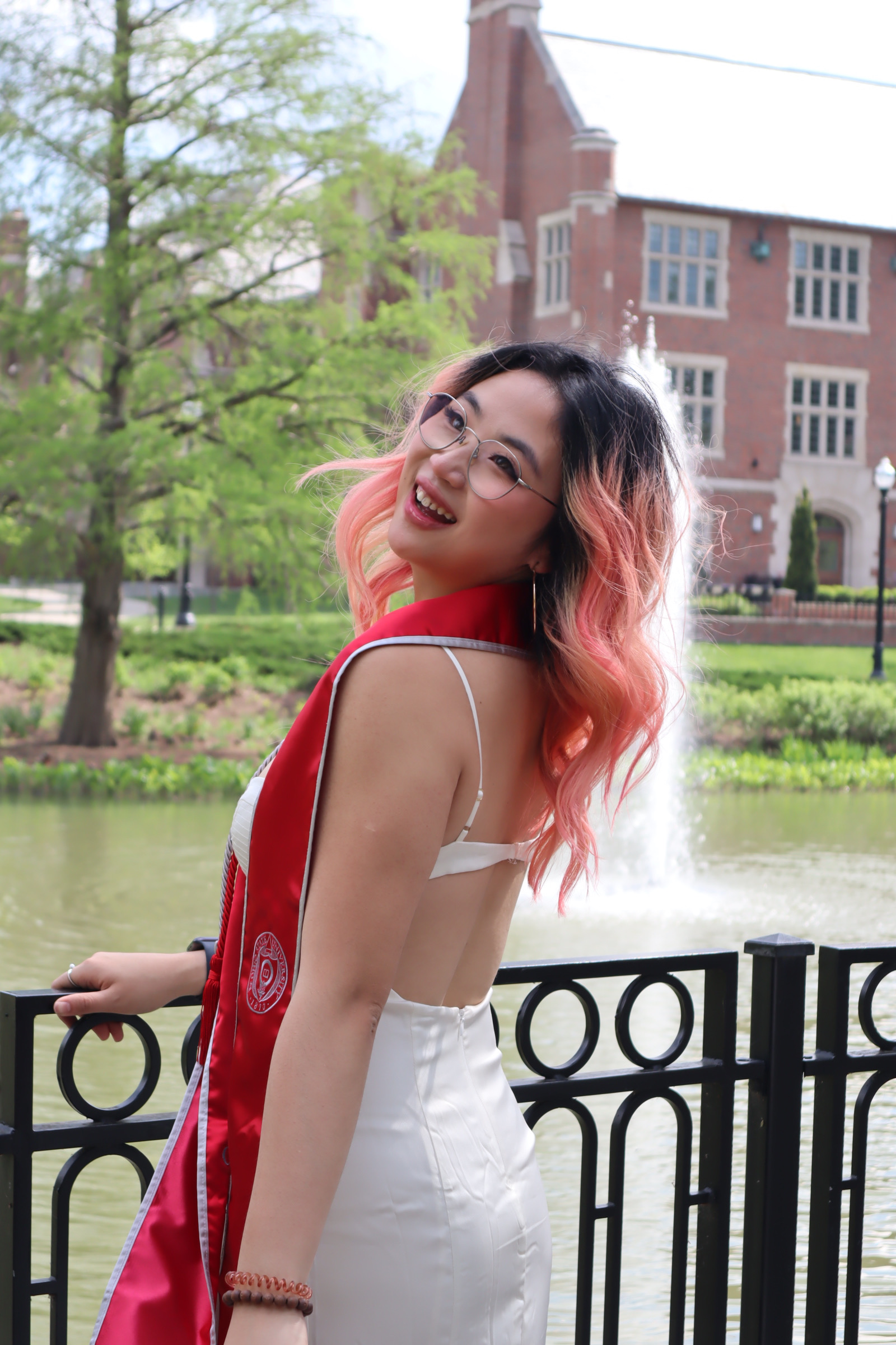 three quarters length picture of a young woman holding a railing and looking over her shoulder, smiling. She has long wavy, red and black hair and is wearing a white dress.