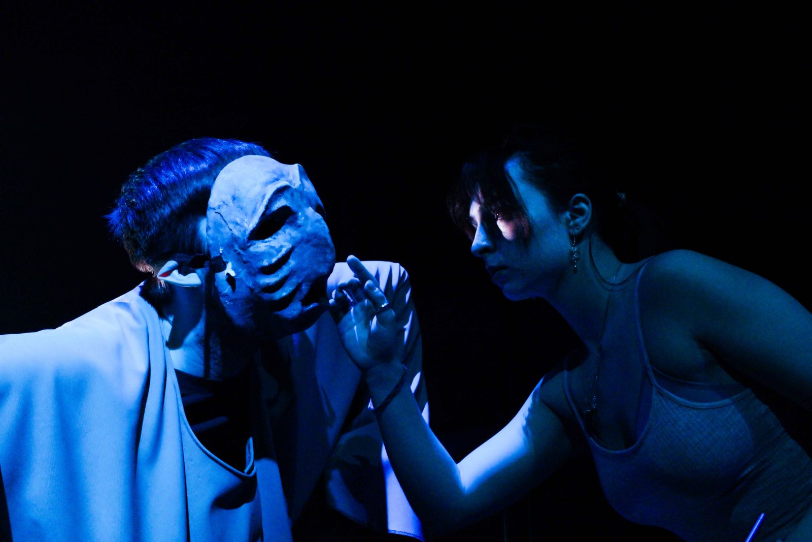 Two actors perform on stage in blue light. The actor on the left is wearing a mask.
