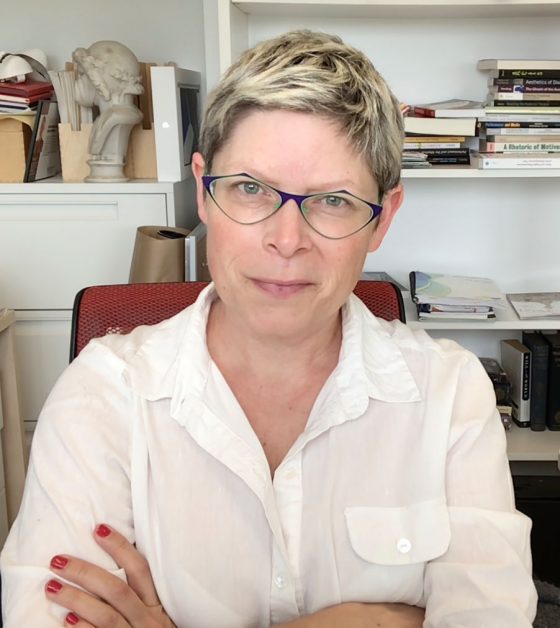 A snapshot of Dr. Jennifer Roberts-Smith (Canadian of Scandinavian-Celtic ethnicity) as she sits in an academic office filled with books. She has a light smile as she looks directly at the camera while wearing striking colourful glasses.