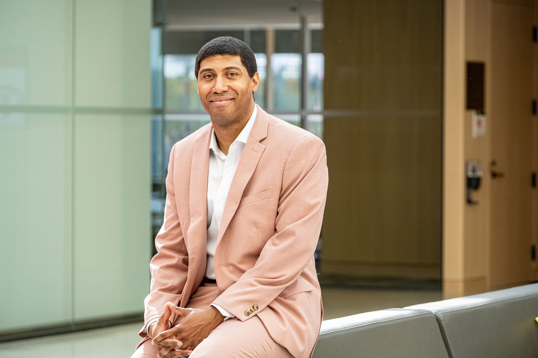 Headshot of a male in a light pink suit sitting on the armrest of a couch