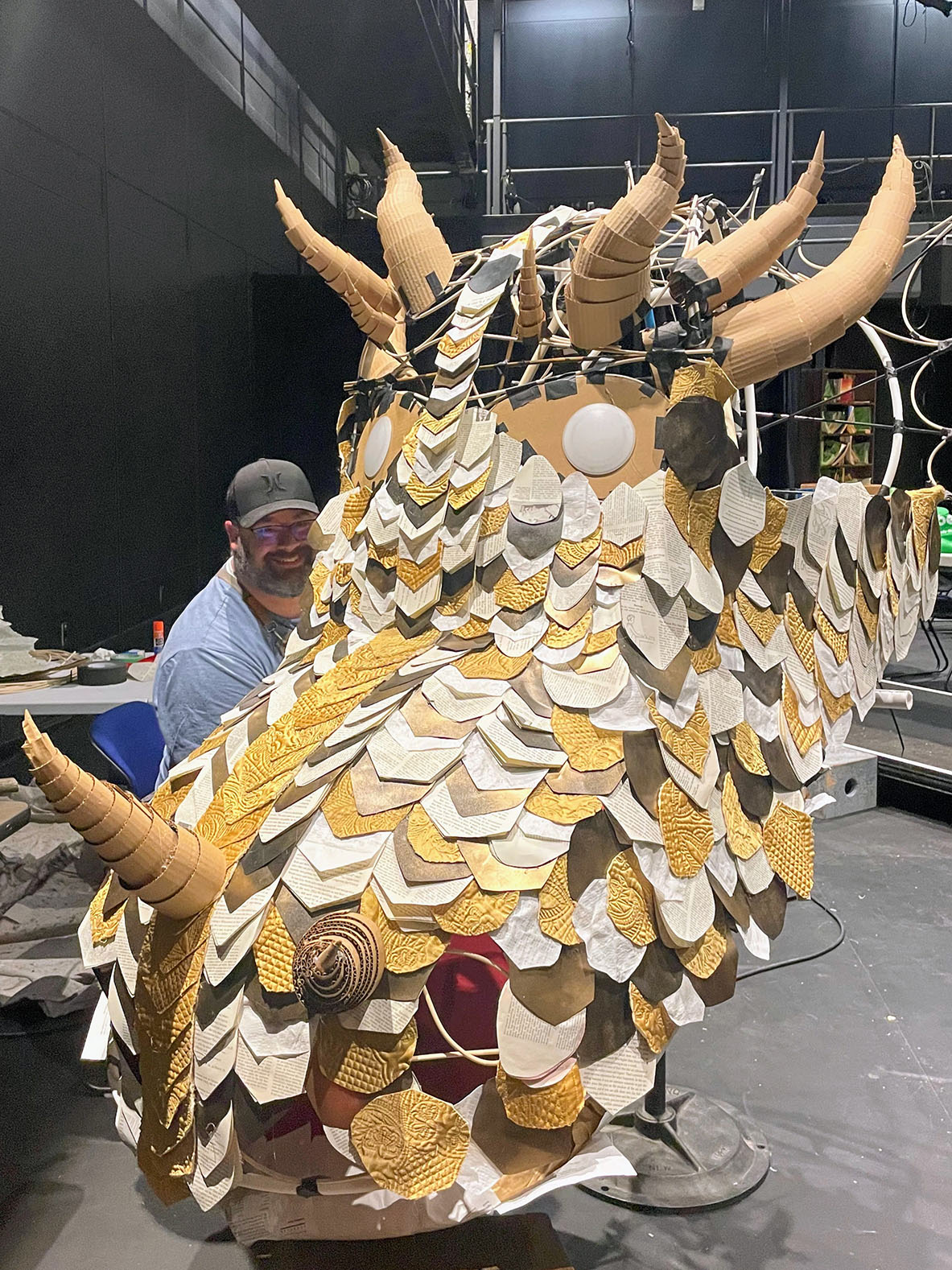A man smiles behind a large, intricately crafted dragon head made of gold, white, and grey scales with ridged cardboard horns.