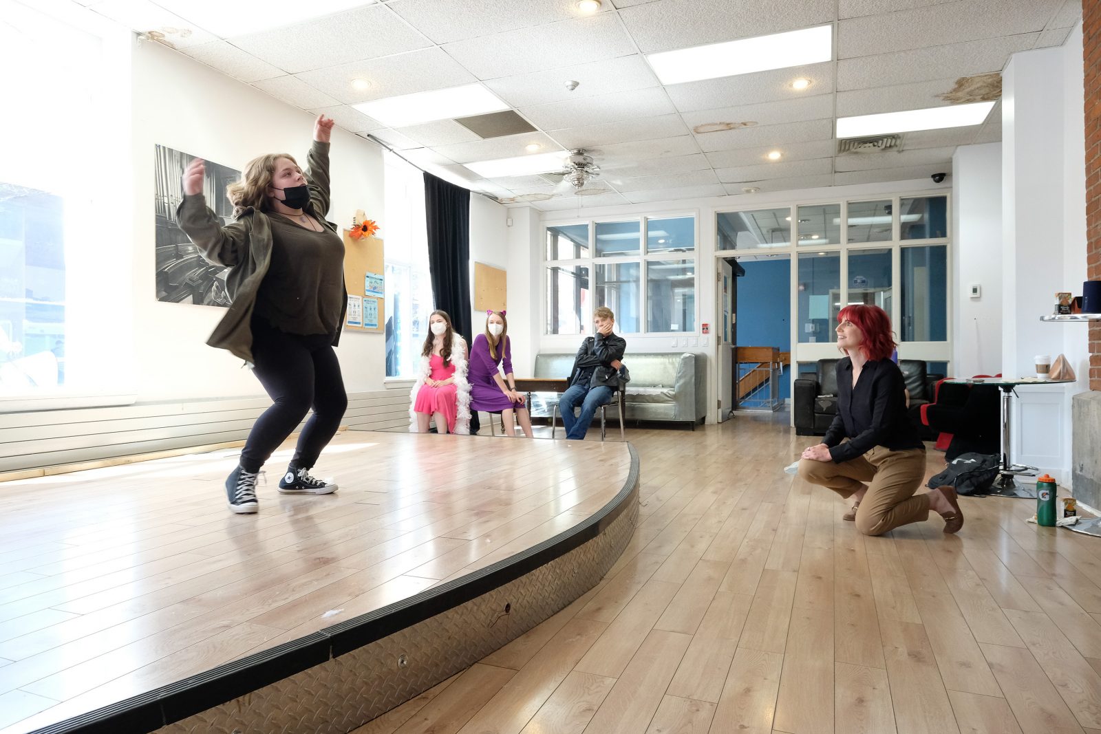 A female performer with her arms up on stage while another person watches kneeled on the ground in front.