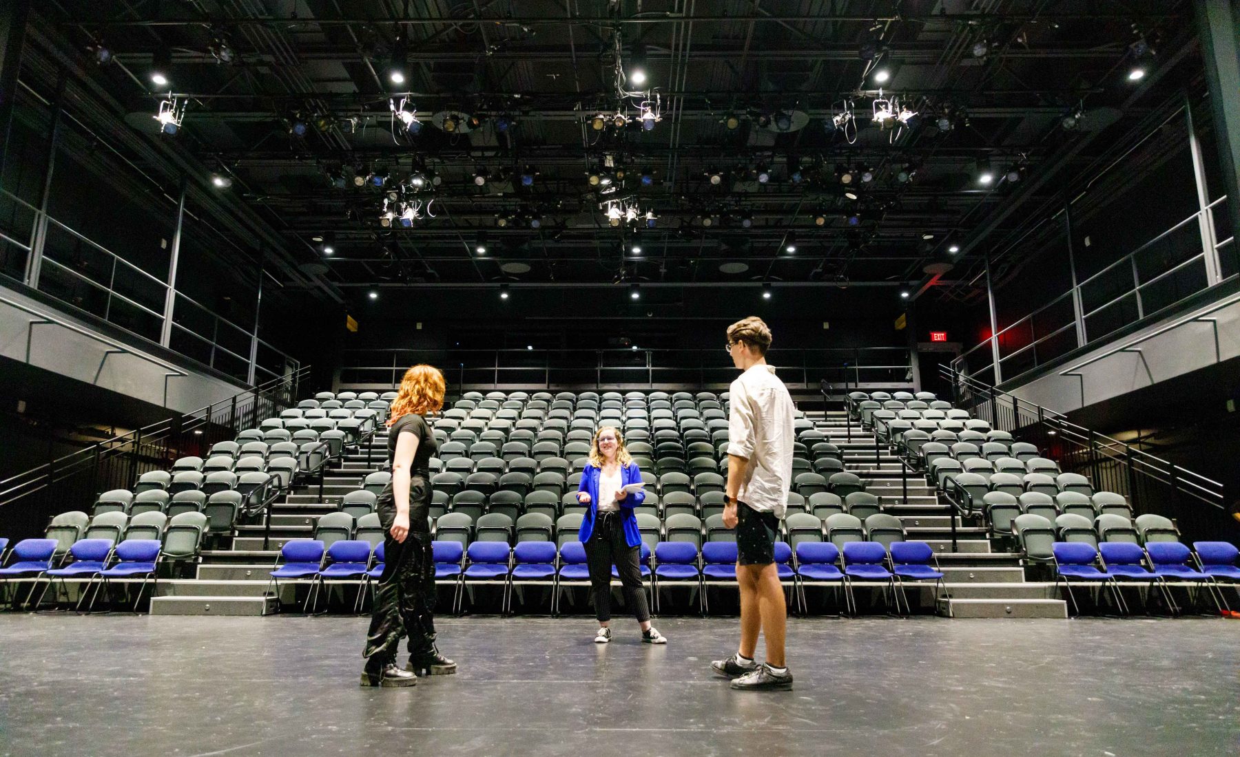 Three people stand on an empty stage with a bank seats and theatre lights behind them. 