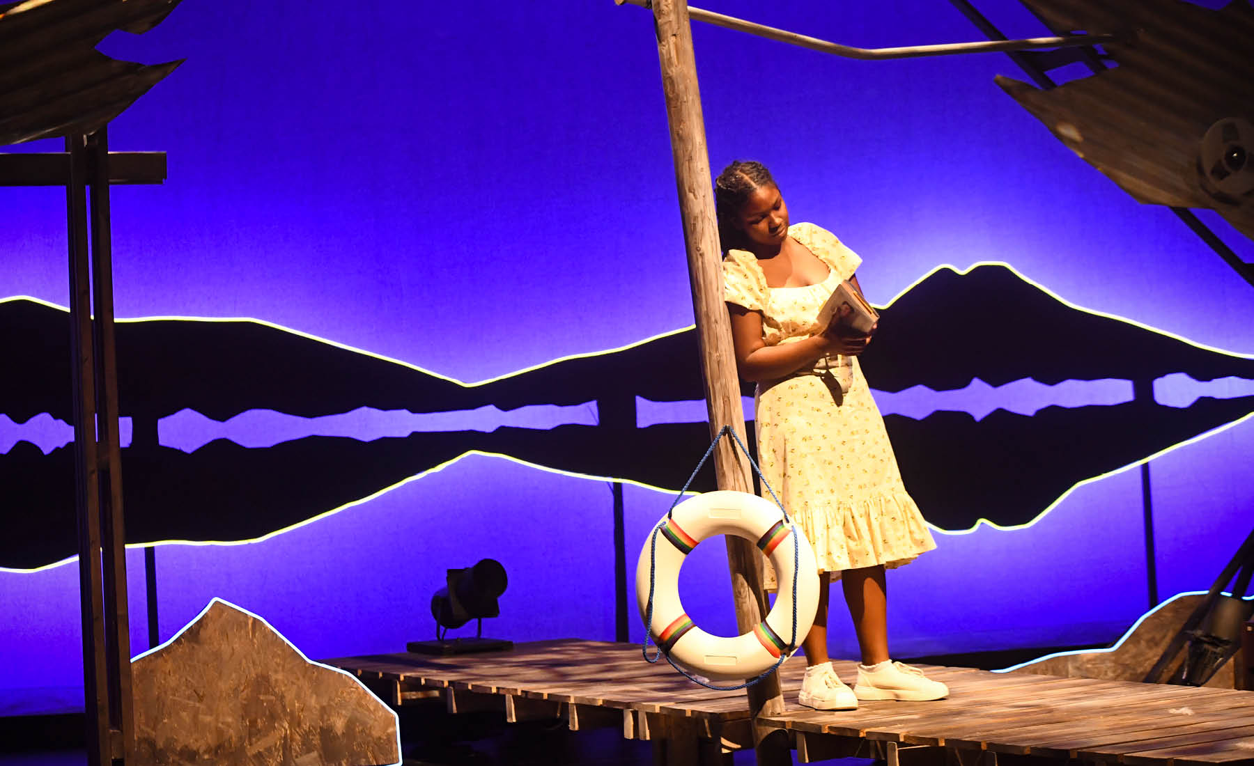 A woman stands on a stage set with a striking blue background, leaning against a post. 