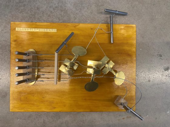 Photo of mixed media art by jef chippewa, brass cut-out new music notation notes entangled with guitar strings affixed onto board.