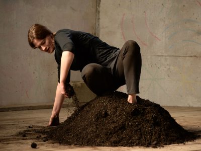 artists on dirt pile wearing black sifting through dirt in an industrial room