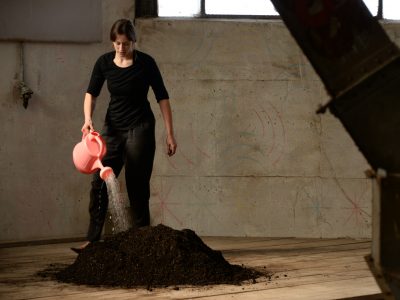 artist holding a pink plastic watering can watering dirt pile in an industrial space