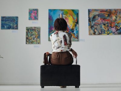 A person with their back towards the camera sits observing five paintings on a gallery wall.